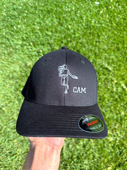 A black baseball cap held up by a hand against a green grass background. The cap features a white embroidered graphic of a person playing disc golf and the text 'CAM' on the front, with a visible Fabri 500 sticker on the brim.